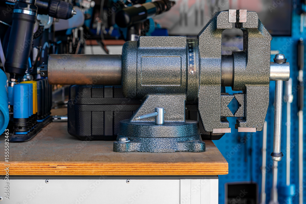 Fototapeta premium Metal vise mounted on a wooden workbench in a workshop. Tools and a blue perforated panel are visible in the background. The image shows sturdy equipment at a mechanic's or DIY workstation
