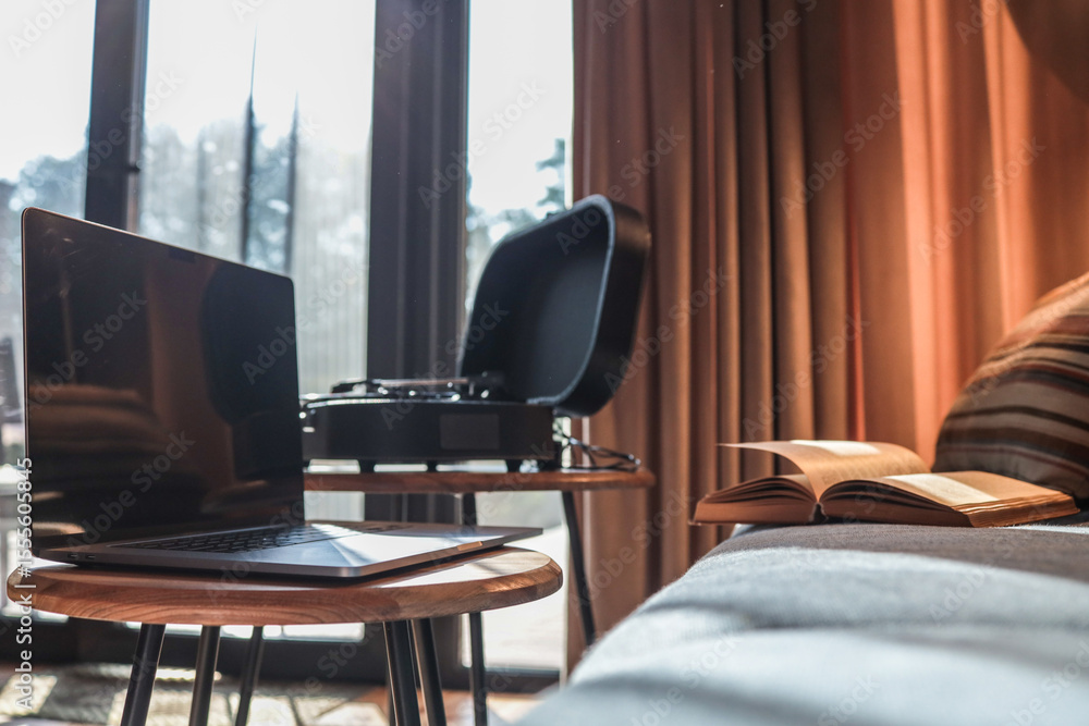 Fototapeta premium Laptop on a small round table with a turntable and open book in the background, set in a cozy interior with warm sunlight and curtains