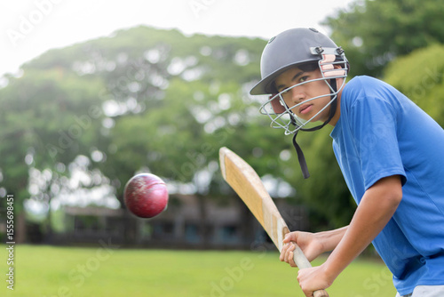 A teenage boy wearing a blue shirt and helmet holding a cricket bat, playing and hitting cricket ball on a school field. He looks focused and ready for action, in motion, selective focus on boy.