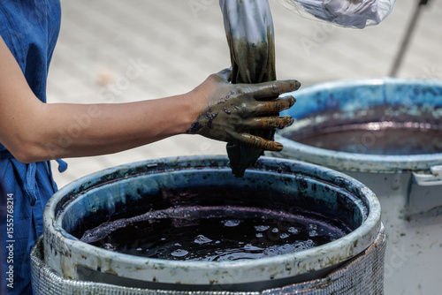 Bean sized indigo blue droplet falls from pinky of artisan's dye soaked hand as it wrings out a fabric's excess wash, returning it to large circular dye vat filled with floating bubbles on surface.