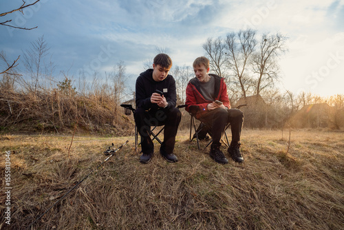Two friends relax outdoors at sunset, looking at smartphones. Fishing gear suggests a fishing trip pause.