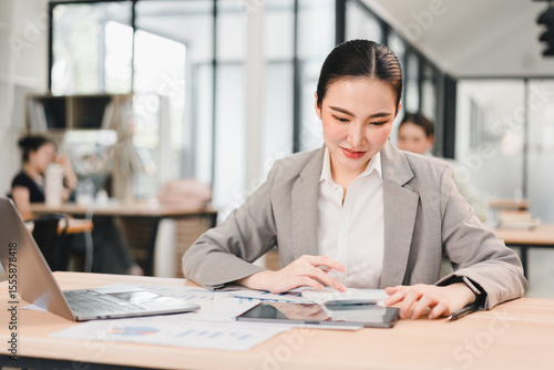 Young businesswoman working with tablet and laptop in modern office, focused and confident expression, casual business attire, bright workspace