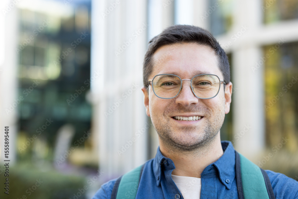Obraz premium A smiling man with glasses is captured outdoors, likely a student or professional, posing in front of a building.