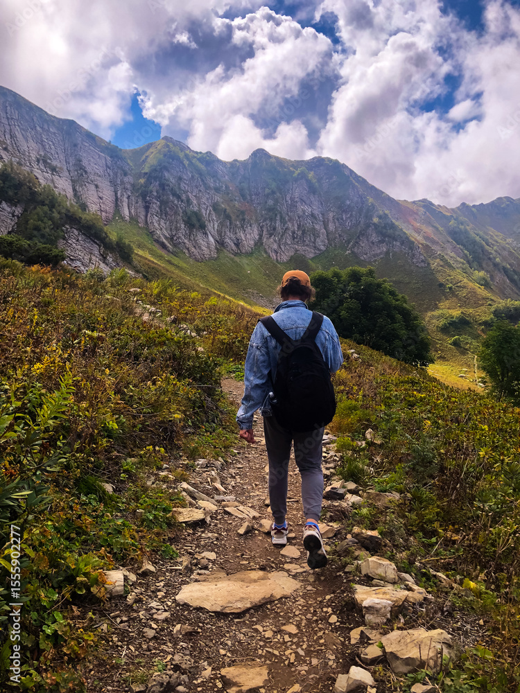 Fototapeta premium Man with a backpack hiking along the Path of Health trail at Rosa Khutor resort in Sochi, Russia, seen from behind. Concept of outdoor adventure, active travel, and exploring alpine landscapes