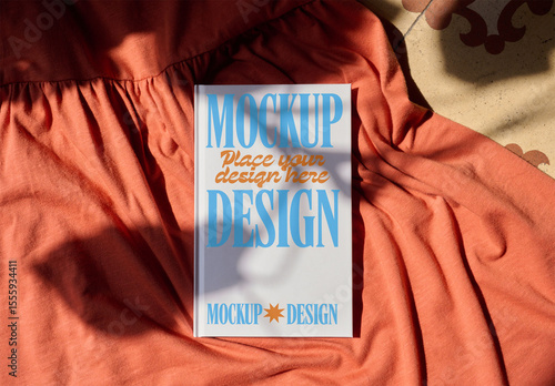 A blank notebook cover rests on soft fabric. Shadows from surrounding objects create interesting patterns on the surface. PSD template mocky¡up