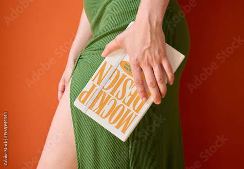 Anonymous Woman dressed in a green dress holds a sleek notebook cover against a bright orange background. PSD template mockup.