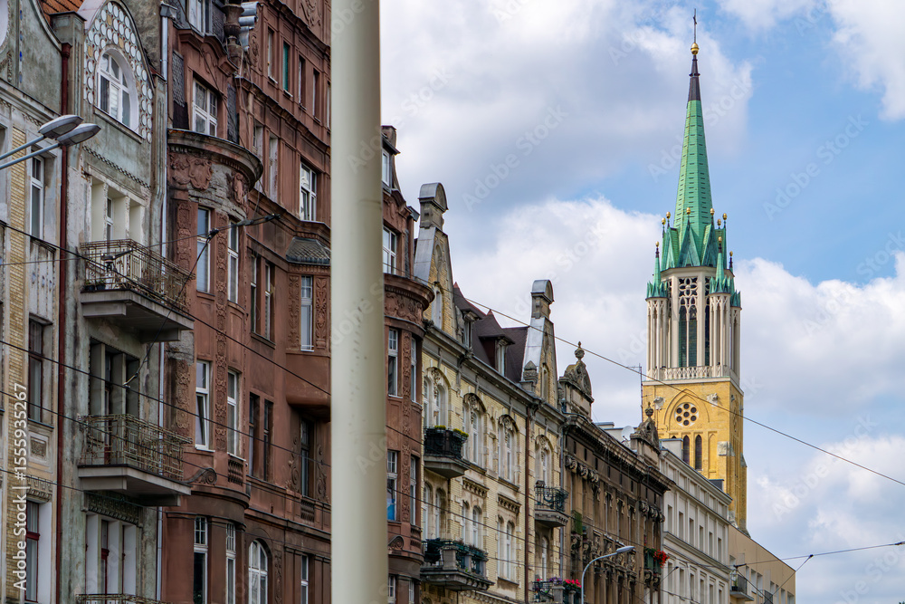 Fototapeta premium Street view in Łódź featuring historic Art Nouveau tenement houses and the tall spire of the St. Stanislaus Kostka Archcathedral Basilica in the background. A blend of classic urban architecture and s