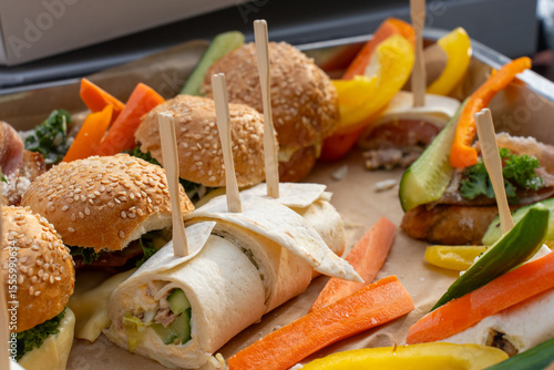 Delicious party platter with sesame seed bagel sandwiches, tortilla wraps and fresh colorful vegetables on wooden board with toothpicks. Healthy catering, appetizer buffet concept. High quality photo