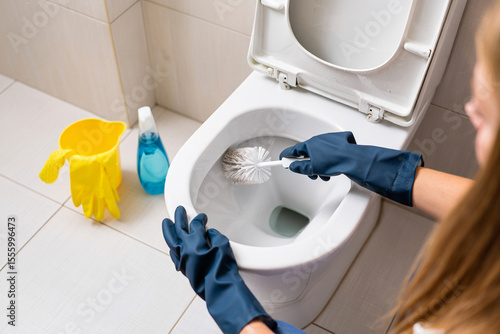 A woman in gloves cleans a toilet bowl with a brush in a brightly lit bathroom.