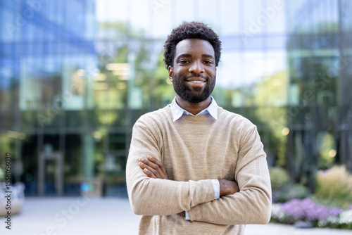 A smiling man with crossed arms stands in front of a modern building. He is dressed casually and exudes confidence.