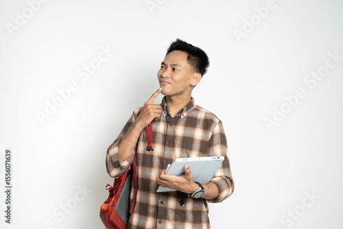 Young student wearing a checkered shirt standing confidently, holding a tablet and a backpack, thinking deeply.