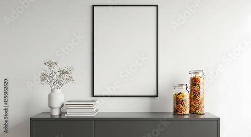 Fototapeta Naklejka Na Ścianę i Meble -  Minimalist interior with blank frame, flowers, books, and pasta jars on a gray cabinet against white wall
