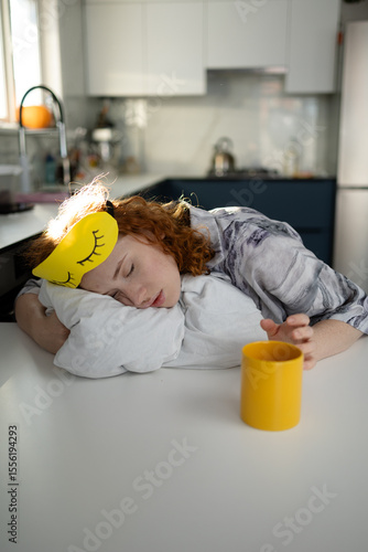 A sleepy, dissatisfied sleep mask girl is sitting in the kitchen, a concept that shows the need for a good night's rest. High quality photo