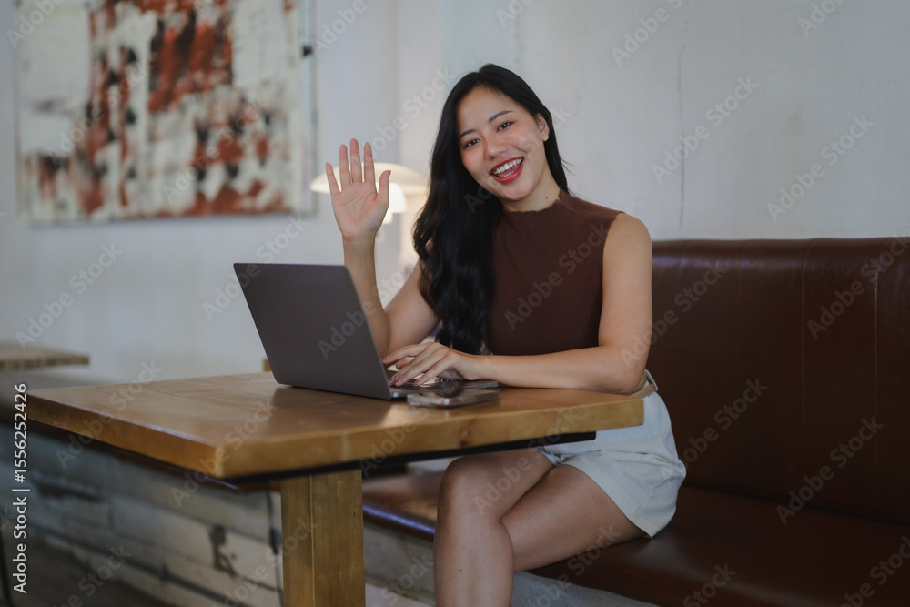 Fototapeta premium Young asian businesswoman waving at laptop during a video call in a cafe