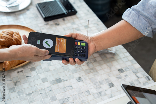 Customer holding smartphone with screen displaying virtual credit card and payment approval while completing cashless transaction on POS terminal held by staff over marble cafe countertop
