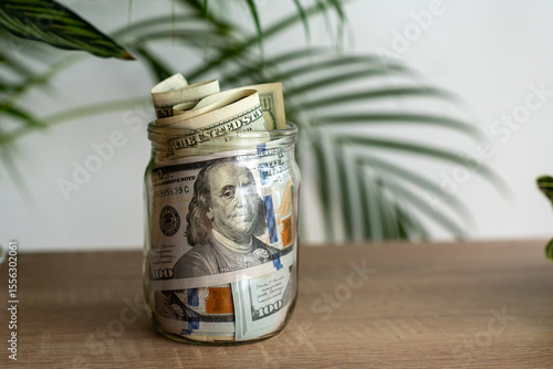 Glass jar full of rolled and stacked US hundred dollar bills on wooden table with green tropical plant in background. Concept of savings, wealth, cash reserve, financial security, and investment.