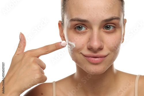 Young woman smiling while applying face cream on acne-prone skin, studio shot on white background