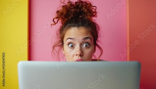 Woman with surprised expression looks over laptop. Colorful background emphasizes amazement. Astonished female face with wide eyes, expressing shock. Modern tech and internet concept.