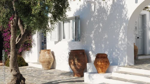 Wallpaper Mural Large vintage terracotta pots near a whitewashed Greek house with open shutters, stone pavement, olive tree shadow, and pink flowers. Shot in Athens, Greece. Sunny day. No people. Torontodigital.ca