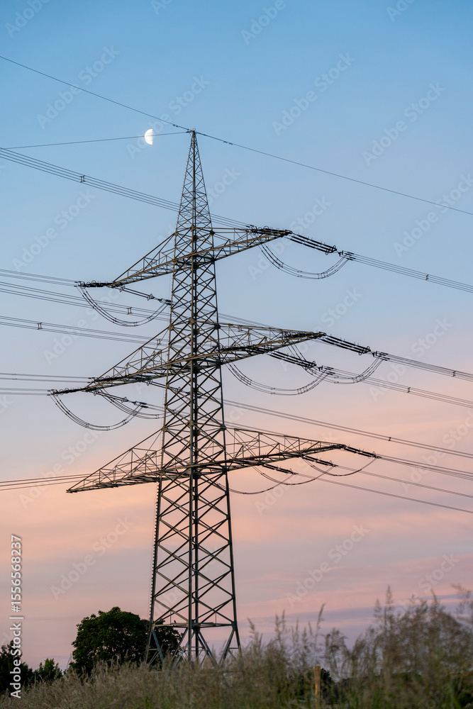 Fototapeta premium A high-voltage transmission pylon stands silhouetted against a vibrant sunset sky with the moon peeking through clouds, blending industrial structure and natural beauty