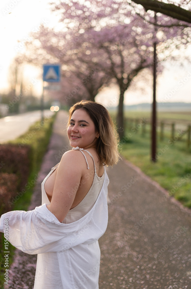 Smiling woman glancing back over her shoulder, on a scenic path lined with pink cherry blossoms. Conveys joy, spontaneity, and natural charm. Smiling woman glancing back over her shoulder, on a scenic path lined with pink cherry blossoms. Conveys joy, spontaneity, and natural charm.