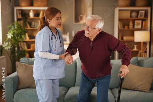 Фототапет A female home care nurse in scrubs assists an elderly man using a cane to stand up from his couch