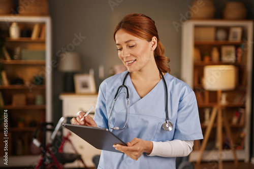 A female healthcare worker, wearing light blue scrubs and a stethoscope, uses a stylus to take notes on a tablet. She is in a living room with bookshelves and warm lighting.