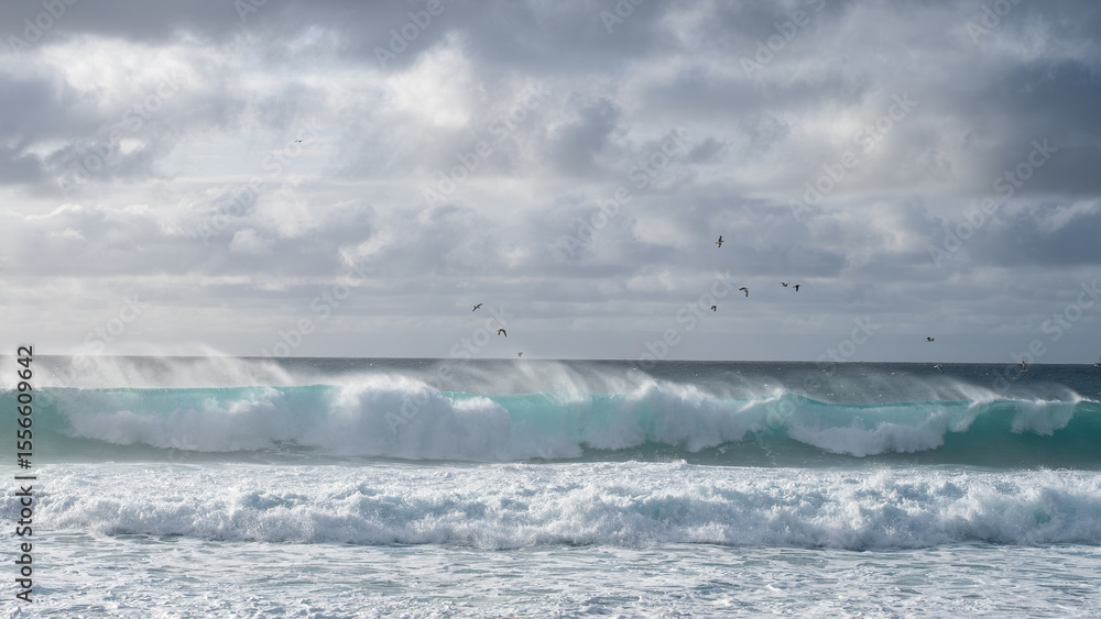 Fototapeta premium Strong wind blows spray off large turquoise waves under a dramatic cloudy sky.
