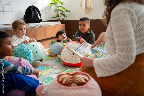 Preschool or kindergarten children sit on the floor listening as the teacher reads a storybook aloud. Concept of early literacy, attention, and storytelling in childhood education