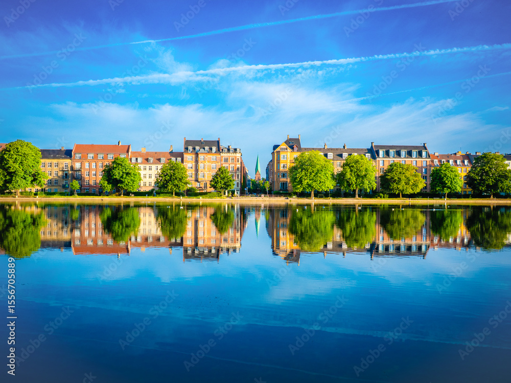 Fototapeta premium Colorful buildings and lush trees reflect on calm waters of Peblinge Sø, Copenhagen, under a vivid blue sky—capturing serene urban beauty in summer.