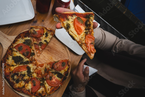 Overhead shot of a person holding a slice of tomato and pesto pizza from a wooden board. Casual dining scene with friends at a cozy café or pizzeria