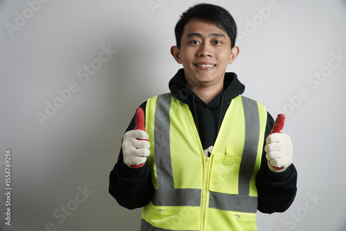 Young Asian Man in Safety Vest Giving Thumbs Up, Asian male worker in a reflective safety vest shows approval or success with a thumbs-up, conveying safety and positivity.