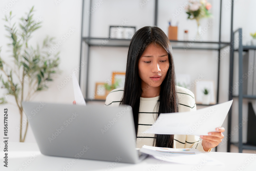 Focused young asian woman working from home, reading and analyzing paperwork with laptop open on desk, multitasking between digital and printed information in modern home workspace Focused young asian woman working from home, reading and analyzing paperwork with laptop open on desk, multitasking between digital and printed information in modern home workspace
