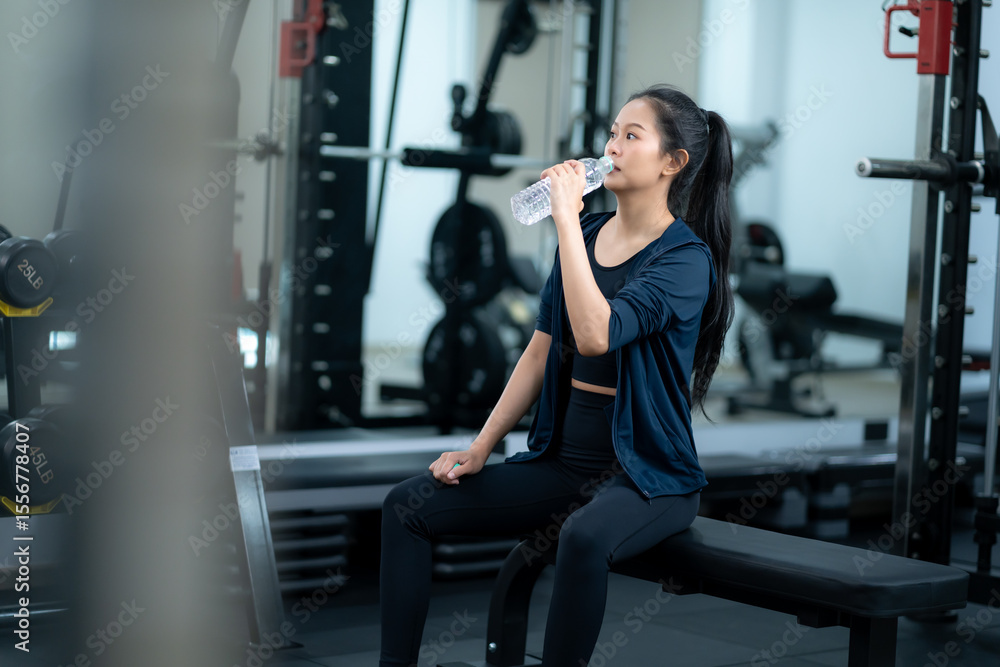 Young asian woman taking a break from workout in gym, sitting on bench with water bottle in hand, wearing sportswear and jacket, showcasing fitness recovery and healthy lifestyle Young asian woman taking a break from workout in gym, sitting on bench with water bottle in hand, wearing sportswear and jacket, showcasing fitness recovery and healthy lifestyle