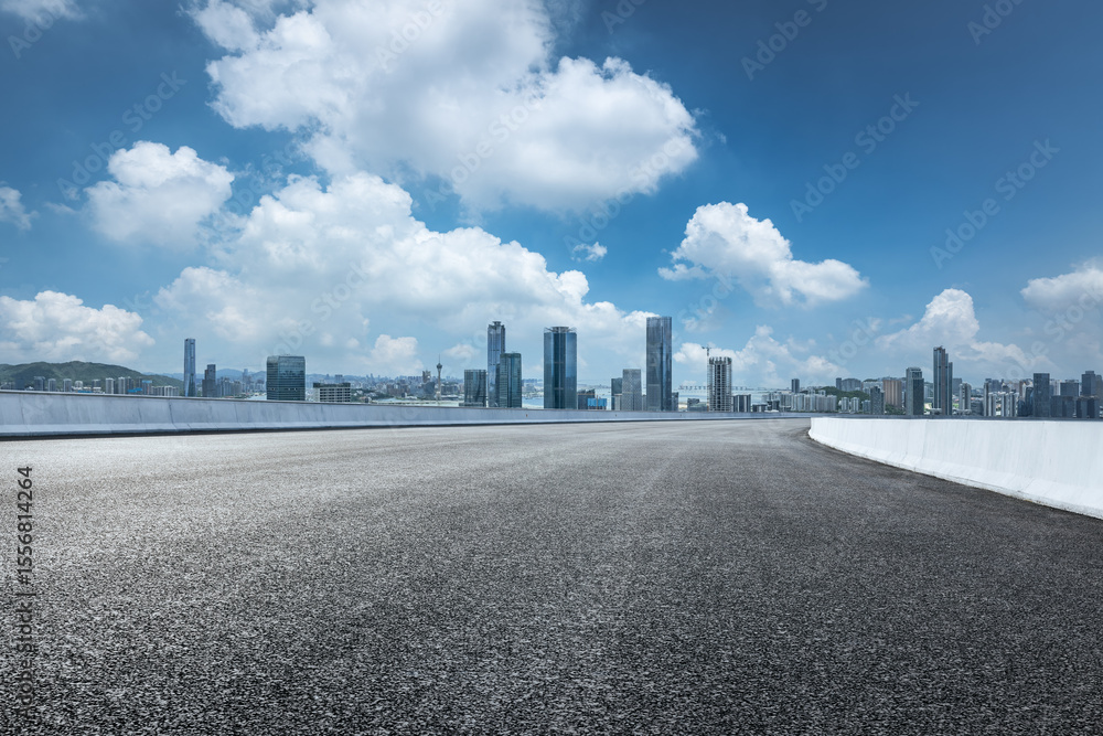 Obraz premium Empty asphalt road with a modern city skyline and commercial buildings in the distance under a beautiful blue sky.