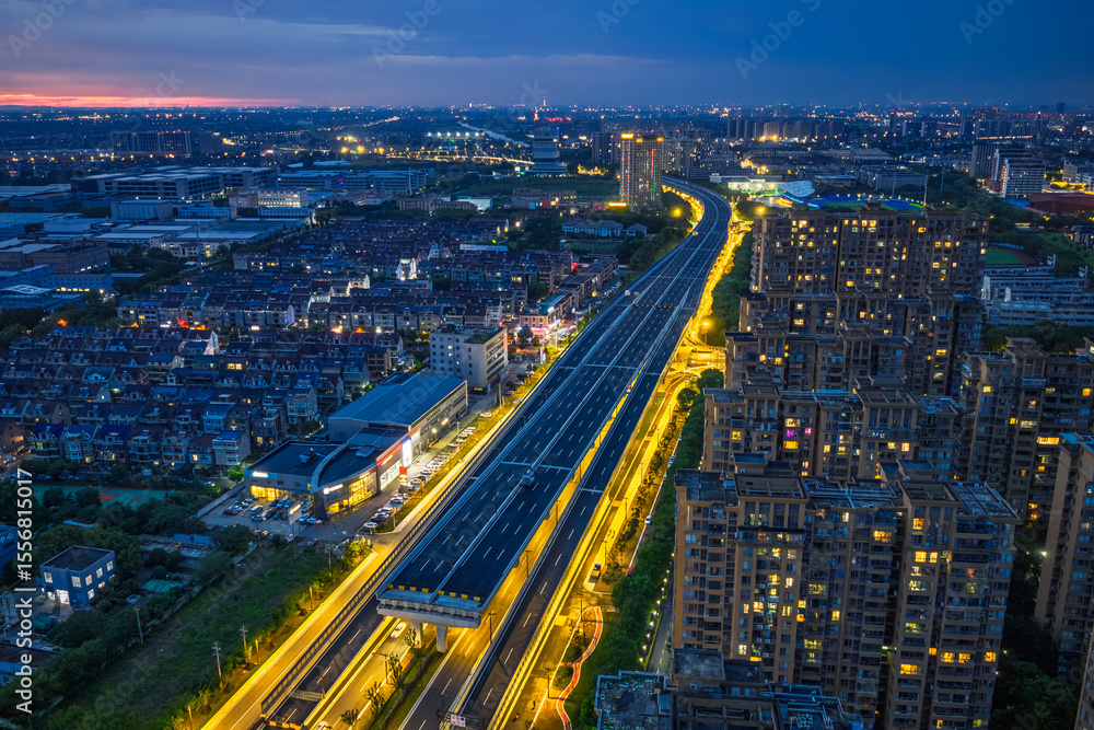 Fototapeta premium Modern elevated expressway illuminated with golden lights cutting through residential and industrial districts at twilight.