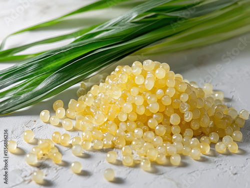 Pile of translucent yellow sago pearls is displayed on textured white surface, accompanied by fresh green pandan leaves, creating vibrant and natural setting
