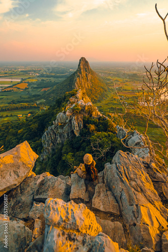 Picture from the back of a woman sitting on the peak of a high mountain cliff. There are a lot of rocks with white. The sun is setting on the mountain, and there is a beautiful warm orange light.