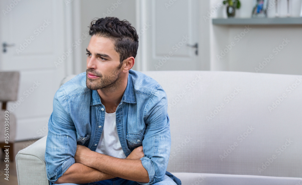 Fototapeta premium Man leaning forward on sofa in living room wearing denim shirt with potted plant, books