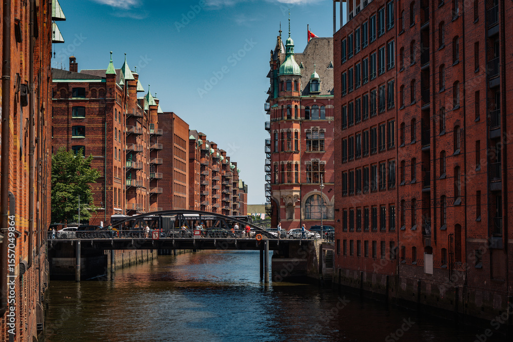 Naklejka premium Canal view through Hamburg’s Speicherstadt reveals pointed green rooftops, ornate facades, and multiple bridges connecting the red-brick buildings.