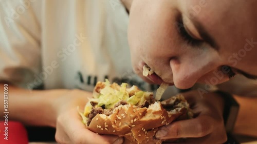 Close-up of a young woman taking a big bite of a sesame seed beef burger in a fast food restaurant. Her expression shows enjoyment and focus on the meal. Concept of fatty and unhealthy food
