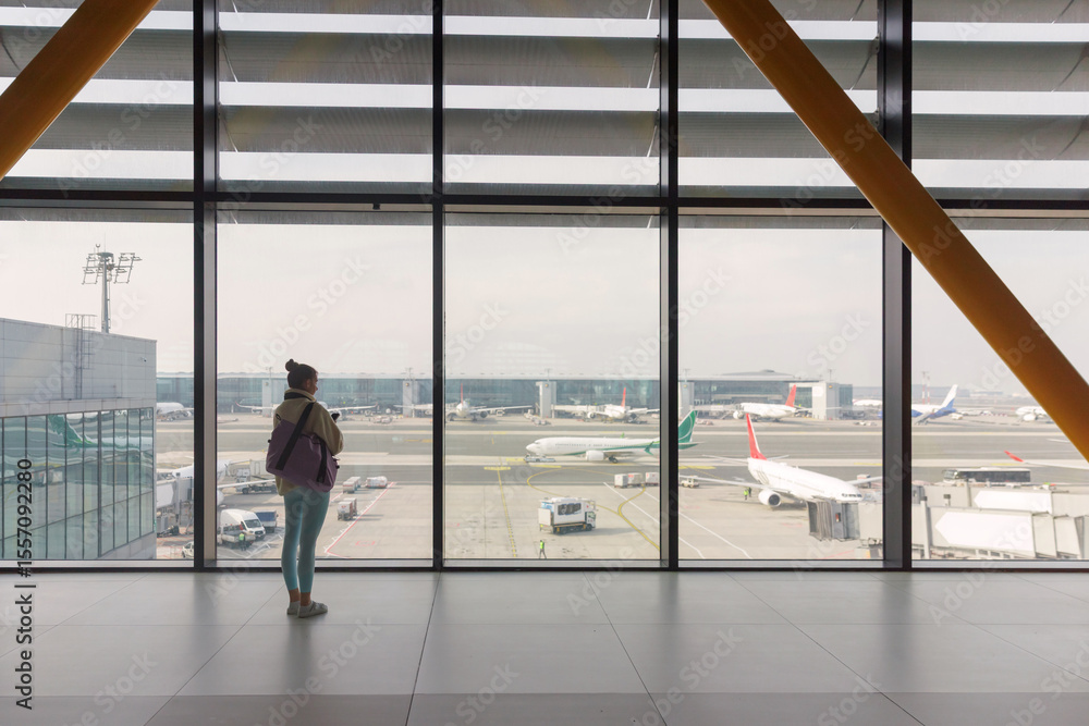 Fototapeta premium A lone traveler stands by a large terminal window, observing aircraft and ground crew activities at a busy airport on a clear day. The scene captures the hustle of air travel.