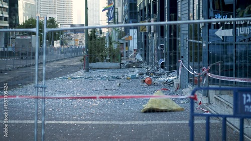 Ground-level side view of a Tel Aviv street filled with broken glass and debris, seen through safety fences after an Iranian missile strike.