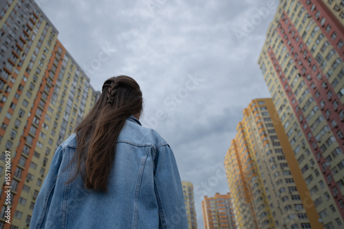 A young woman stands with her back to the camera looking up at tall, brightly colored apartment buildings against a dramatic sky. The atmosphere is reflective and urban