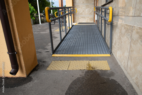 A well-constructed accessibility ramp made of metal features yellow handrails and is located at the entrance of a public building. The pathway is clean and mottled with wet patches