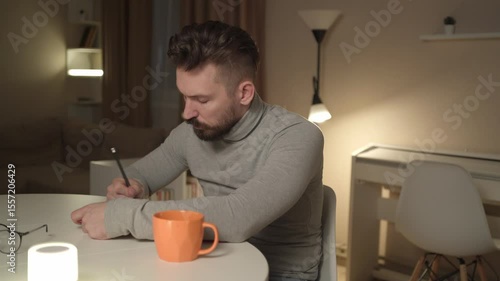 Bearded 30-year-old man sits in the living room in the evening, reading his daily planner and writing down ideas and thoughts. A natural, unaltered moment of focus and reflection in a cozy atmosphere