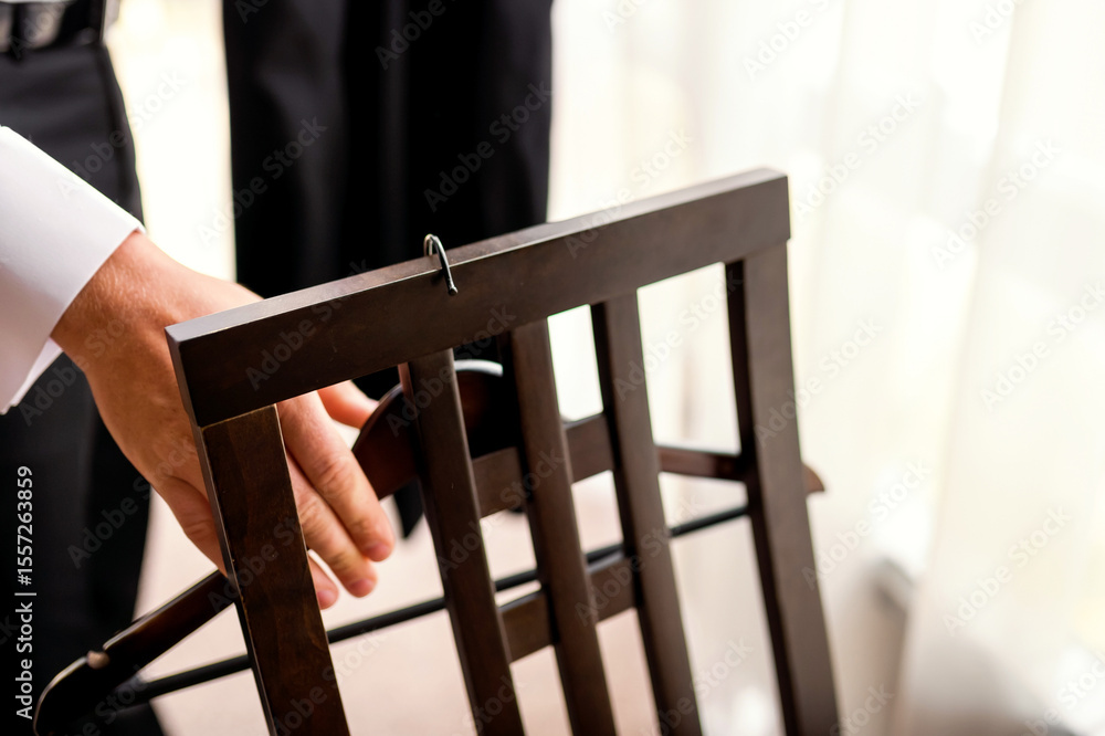 Obraz premium Hand grasping a wooden chair. A hand grips the back of a dark wooden chair in a well-lit indoor space, suggesting preparation for an event.