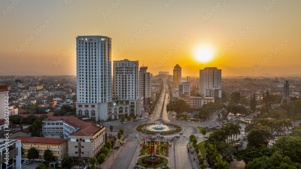 Fototapeta premium The sun sets or rises behind buildings in Thai Nguyen city, casting a warm glow over the central roundabout and urban area.