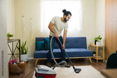 Фотография Latin man vacuuming carpet in living room doing housework
