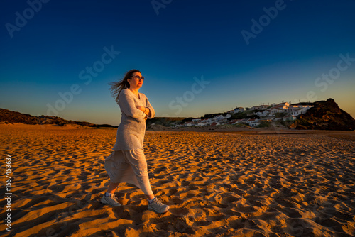 Beautiful mid adult woman walking on sandy beach on sunset on windy spring day. Side view. Monte Clerigo beach on Algarve coast in Portugal	